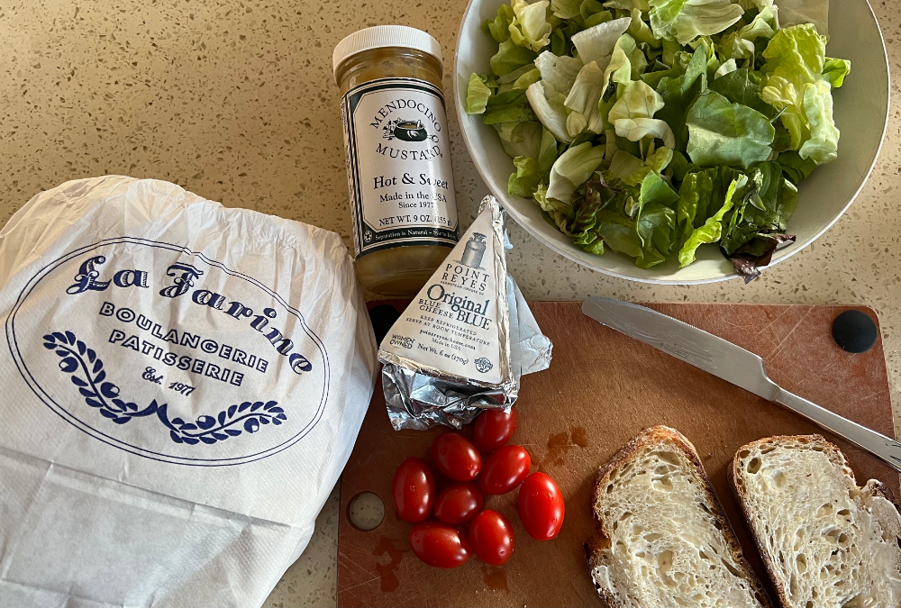 Photograph of K's sandwich ingredients on the kitchen counter: jar of mustard, wedge of blue cheese, bowl of lettuce, handful of little tomatoes, two slices of bread and the rest of the load in its bakery bag.